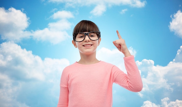 concept d'enfance, d'école, d'éducation, de vision et de personnes - petite fille heureuse dans des lunettes pointant le doigt vers le haut sur fond de ciel bleu et de nuages