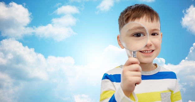 concept d'enfance, d'enquête, de découverte, de vision et de personnes - petit garçon heureux regardant à travers une loupe sur fond bleu ciel et nuages