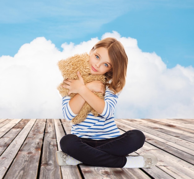 concept d'enfance, de jouets et de personnes - jolie petite fille embrassant un ours en peluche sur fond bleu ciel et nuage