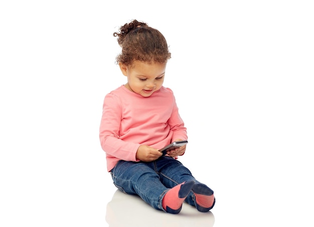 concept d'enfance, de technologie et de personnes - petite fille afro-américaine souriante jouant avec un smartphone et assise sur le sol