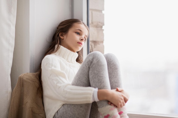 concept d'enfance, de tristesse et de personnes - triste belle fille en pull assis sur le rebord de la fenêtre de la maison en hiver