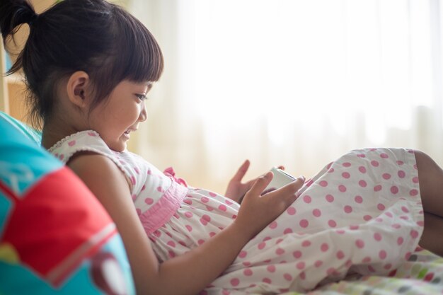 Concept enfant, technologie et internet - petite fille au lit avec smartphone à la maison