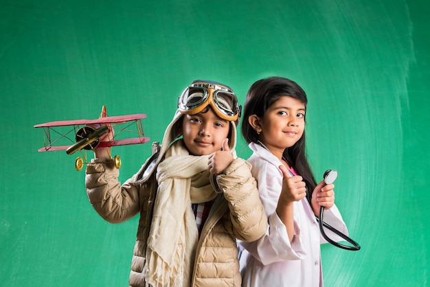 Concept d'enfants et d'éducation - Petit garçon et fille indiens posant devant un tableau vert en déguisement pilote et costume de médecin avec stéthoscope, debout sur fond de tableau vert