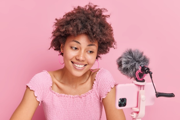 Concept d'enregistrement de podcast et de streaming vidéo de blogs. Une femme afro-américaine souriante a des cheveux bouclés naturels regarde l'appareil photo du smartphone