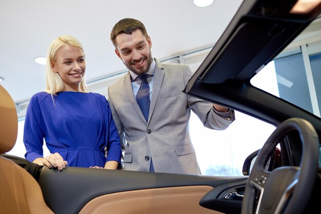 concept d'entreprise automobile, de vente de voitures, de consommation et de personnes - couple heureux achetant une voiture dans un salon de l'auto ou un salon