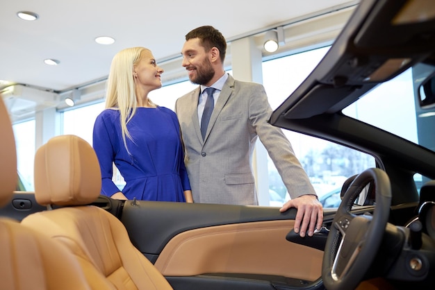 concept d'entreprise automobile, de vente de voitures, de consommation et de personnes - couple heureux achetant une voiture dans un salon de l'auto ou un salon