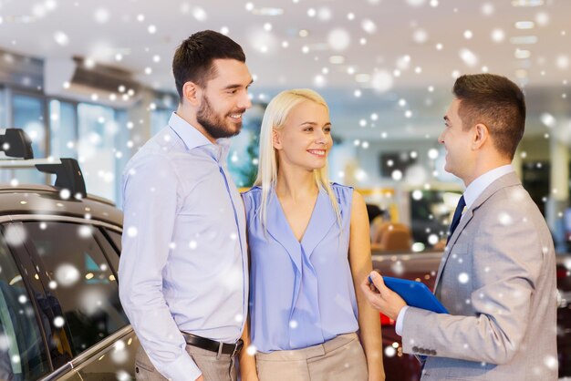 concept d'entreprise automobile, de vente de voitures, de technologie et de personnes - couple heureux avec un concessionnaire automobile dans un salon de l'auto ou un salon sur l'effet de neige