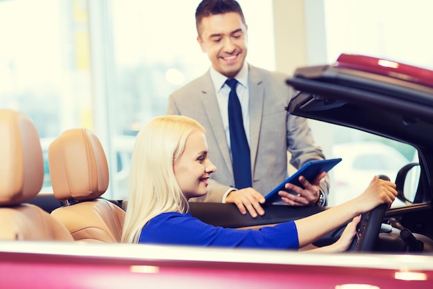 concept d'entreprise automobile, de vente de voitures, de technologie et de personnes - femme heureuse et concessionnaire automobile avec ordinateur tablette dans un salon de l'auto ou un salon