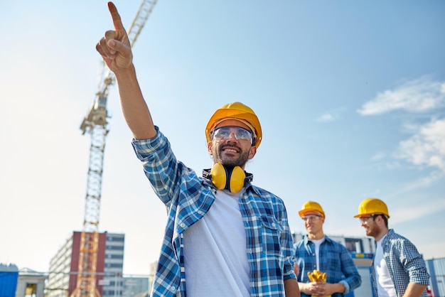 concept d'entreprise, de construction, de travail d'équipe et de personnes - groupe de constructeurs souriants dans des casques pointant le doigt de côté sur le chantier de construction