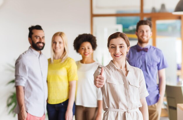 concept d'entreprise, de démarrage, de personnes, de gestes et de travail d'équipe - jeune femme heureuse montrant les pouces vers le haut sur l'équipe créative au bureau