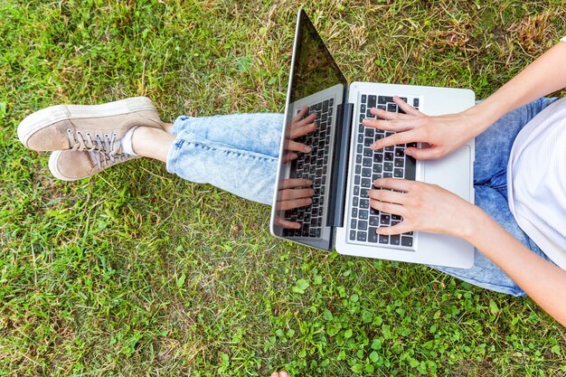 Concept d'entreprise indépendant. Jambes de femme sur la pelouse d'herbe verte dans le parc de la ville, mains travaillant sur un ordinateur portable. Style de vie authentique étudiante candide étudiant à l'extérieur. Bureau mobile