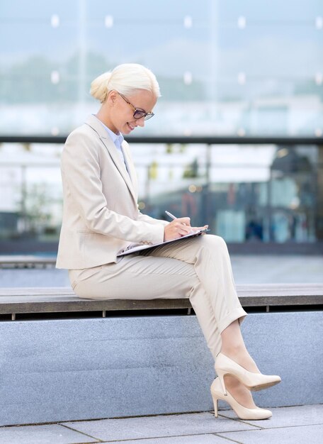 concept d'entreprise, de personnes et d'éducation - jeune femme d'affaires souriante dans des verres avec un bloc-notes sur un immeuble de bureaux