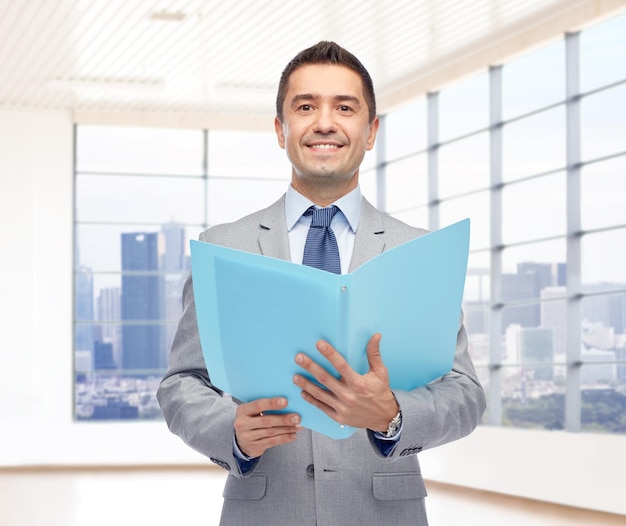 concept d'entreprise, de personnes, de finances et d'immobilier - heureux homme d'affaires souriant en costume tenant un dossier ouvert au-dessus d'une salle de bureau avec une grande fenêtre et un fond de vue sur la ville