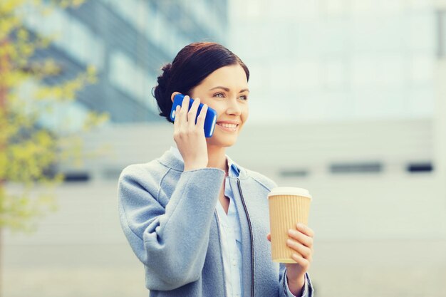 concept d'entreprise et de personnes - jeune femme souriante appelant sur un smartphone et buvant du café dans un immeuble de bureaux en ville