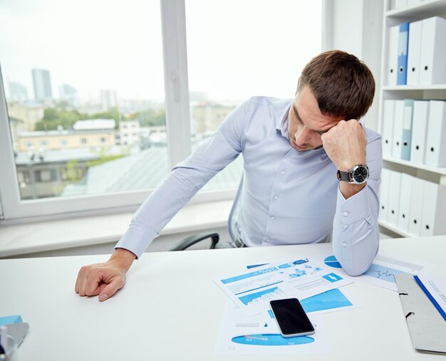 concept d'entreprise, de personnes, de paperasse et de délai - homme d'affaires stressé avec smartphone, papiers et graphiques assis à table au bureau