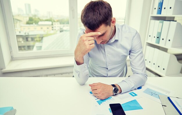 concept d'entreprise, de personnes, de paperasse et de délai - homme d'affaires stressé avec smartphone, papiers et graphiques assis à table au bureau