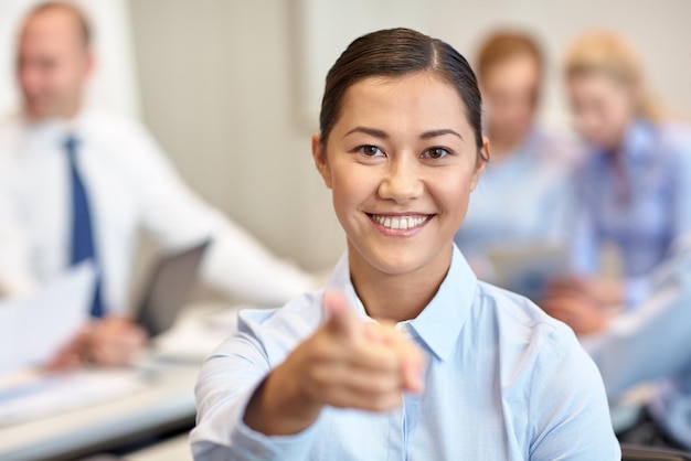 concept d'entreprise, de personnes et de travail d'équipe - femme d'affaires souriante pointant le doigt vers vous avec un groupe d'hommes d'affaires réunis au bureau