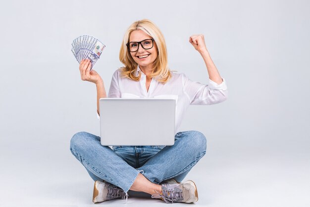 Concept d'entreprise. Portrait d'une femme âgée heureuse en tenue décontractée assise sur le sol en position du lotus et tenant un ordinateur portable et des billets d'argent isolés sur fond blanc. Copiez l'espace pour le texte.