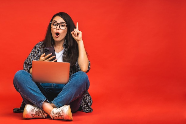 Concept d'entreprise Portrait d'une femme brune étonnée choquée en tenue décontractée assise sur le sol en pose de lotus et tenant un ordinateur portable isolé sur fond rouge à l'aide d'un téléphone portable