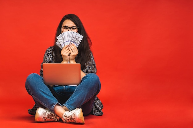 Concept d'entreprise Portrait d'une femme brune heureuse en tenue décontractée assise sur le sol en pose de lotus et tenant un ordinateur portable isolé sur fond rouge