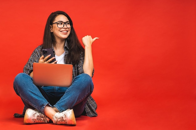 Concept d'entreprise Portrait d'une femme brune souriante heureuse en tenue décontractée assise sur le sol en pose de lotus et tenant un ordinateur portable isolé sur fond rouge à l'aide d'un téléphone portable