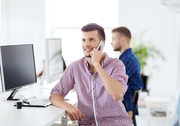 concept d'entreprise, de technologie, d'éducation et de personnes - heureux jeune homme créatif ou étudiant avec ordinateur au bureau appelant sur smartphone