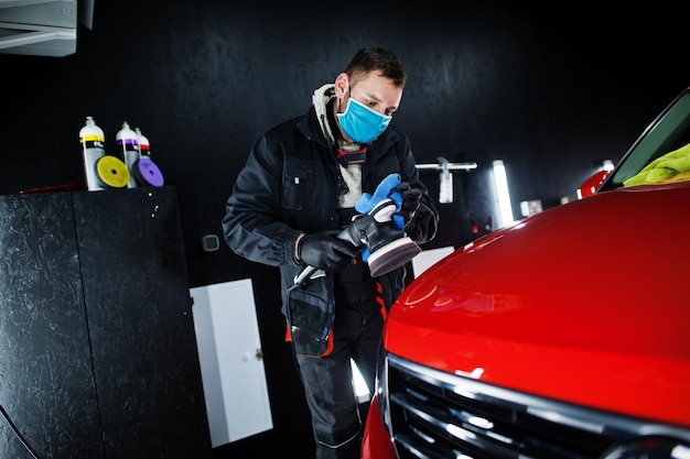 Concept d'esthétique automobile Homme en masque facial avec polisseuse orbitale dans un atelier de réparation polissant une voiture suv orange