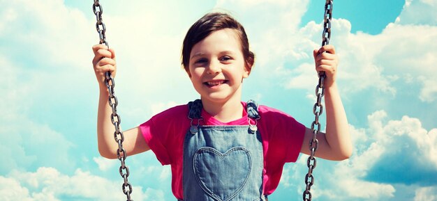 concept d'été, d'enfance, de loisirs, d'amitié et de personnes - petite fille heureuse se balançant sur fond de ciel bleu et nuages