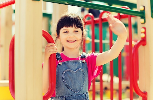 concept d'été, d'enfance, de loisirs, de gestes et de personnes - petite fille heureuse agitant la main sur le cadre d'escalade de l'aire de jeux pour enfants