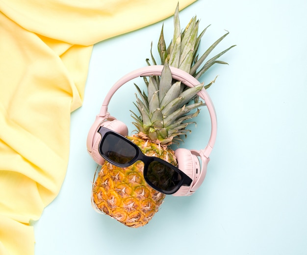 Concept d'été avec fruit d'ananas tropical dans des lunettes de soleil et des écouteurs sur un espace bleu.