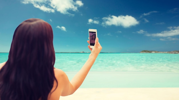 concept d'été, de voyage, de technologie et de personnes - gros plan d'une jeune femme sexy prenant un selfie avec un smartphone sur fond de plage tropicale