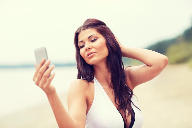 concept d'été, de voyage, de technologie et de personnes - jeune femme sexy prenant selfie avec smartphone sur la plage