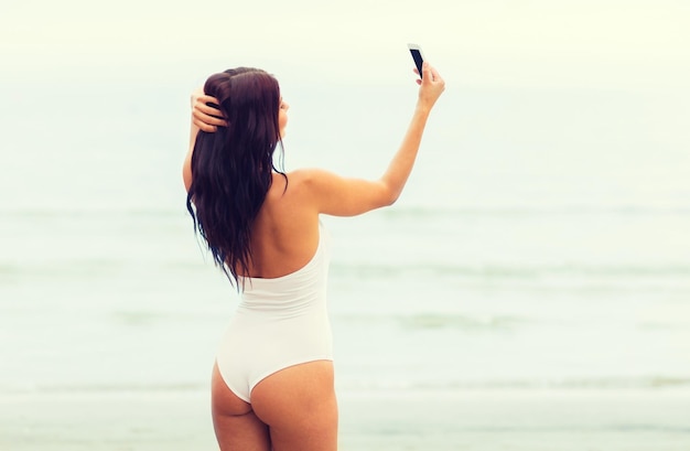 concept d'été, de voyage, de technologie et de personnes - jeune femme sexy prenant selfie avec smartphone sur la plage