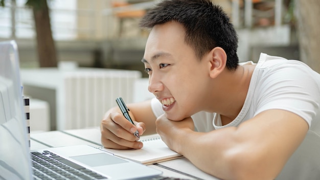 Concept d'étude en ligne un étudiant en t-shirt blanc appréciant d'étudier en ligne et assis devant son nouvel ordinateur portable blanc à l'extérieur.