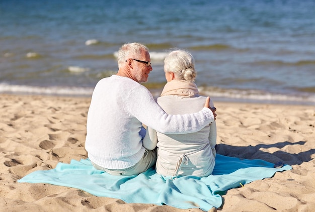 concept de famille, d'âge, de voyage, de tourisme et de personnes - couple de personnes âgées heureux assis sur un plaid et étreignant sur la plage d'été