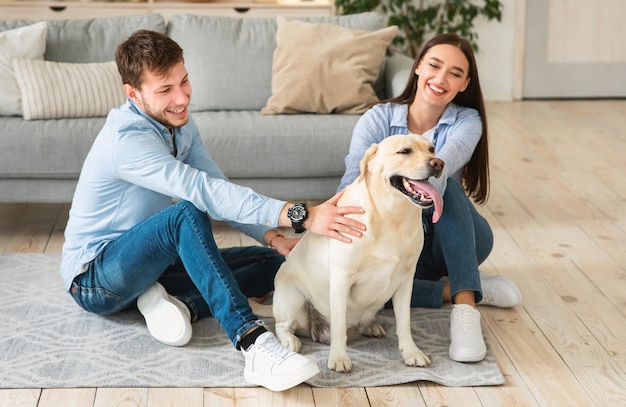 Concept de famille aimante heureuse. Portrait de conjoints joyeux assis par terre dans un appartement moderne, passant du temps avec un chien. Propriétaires d'homme et de femme jouant avec l'animal familier de labrador à la maison