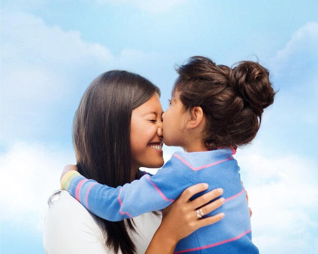 concept de famille, d'enfants et de personnes heureuses - petite fille heureuse étreignant et embrassant sa mère sur fond de ciel bleu