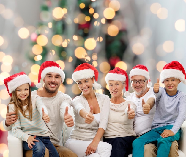 concept de famille, de génération, de geste, de vacances et de personnes - famille heureuse en chapeaux d'assistant de santa montrant les pouces vers le haut sur fond de lumières de sapin de noël