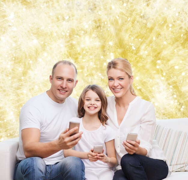 concept de famille, de vacances, de technologie et de personnes - mère souriante, père et petite fille avec des smartphones sur fond de lumières jaunes