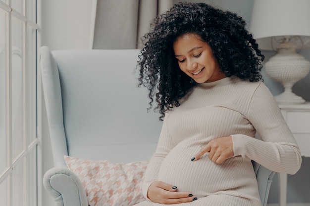 Concept de femmes et de grossesse. Belle femme enceinte heureuse en robe blanche aux cheveux bouclés assise sur un fauteuil confortable à la maison près de la fenêtre et pointant son futur bébé dans son ventre