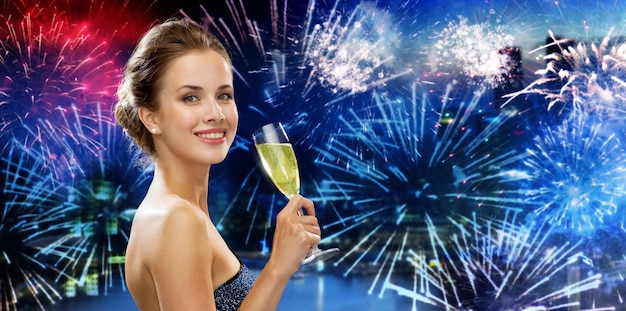 concept de fête, de boissons, de vacances, de luxe et de célébration - femme souriante en robe de soirée avec un verre de vin mousseux sur fond de ville et de feu d'artifice