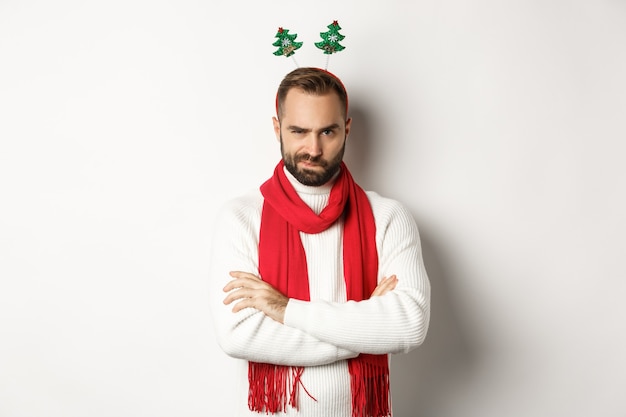 Concept de fête et de célébration de Noël. Jeune homme barbu suspect à l'air douteux, portant un chapeau accessoire drôle, debout sur fond blanc.