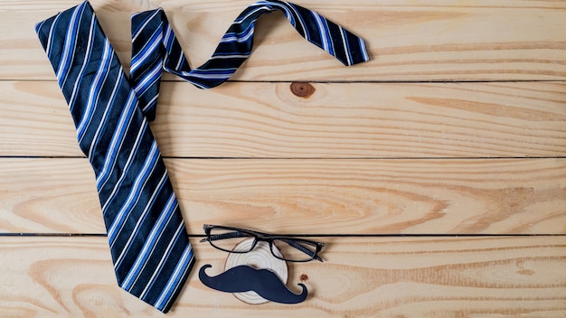 Concept de fête des pères heureux. Un papier à moustache noir, une cravate bleue et des lunettes sur une table en bois.