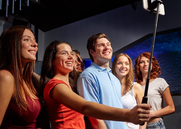 concept de fête, de technologie, de vie nocturne et de personnes - amis souriants avec smartphone et monopode prenant selfie en club