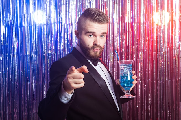 Concept de fête et de vacances de club - Portrait d'un bel homme élégant dans un costume cher détient un cocktail bleu sur fond brillant.