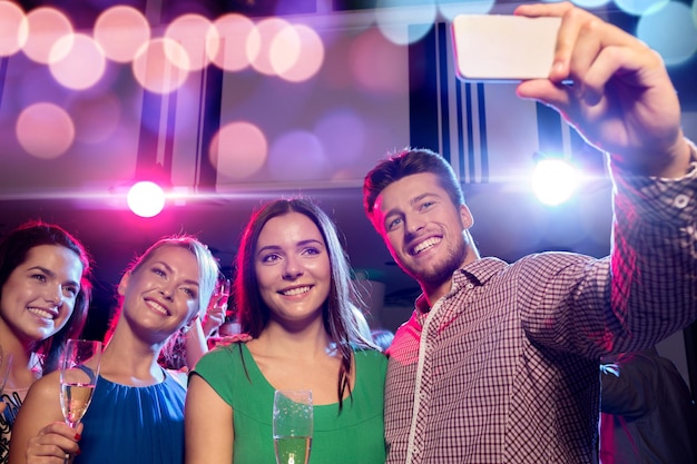 Concept de fête, vacances, technologie, vie nocturne et personnes - amis souriants avec des verres de champagne et smartphone prenant selfie en club