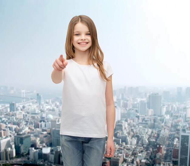 concept de gestes et de personnes heureuses - petite fille heureuse en t-shirt blanc vierge pointant vers vous