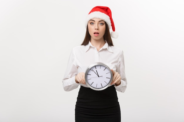 Concept de gestion du temps - jeune femme d'affaires avec bonnet de Noel tenant une horloge isolée sur un mur blanc.