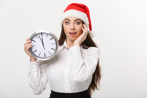 Concept de gestion du temps - jeune femme d'affaires avec bonnet de Noel tenant une horloge isolée sur un mur blanc.