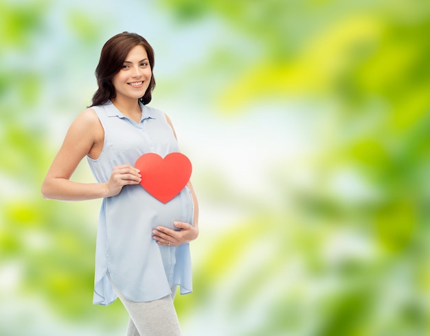 concept de grossesse, d'amour, de personnes et d'attentes - femme enceinte heureuse avec une forme de coeur rouge touchant son ventre sur fond naturel vert